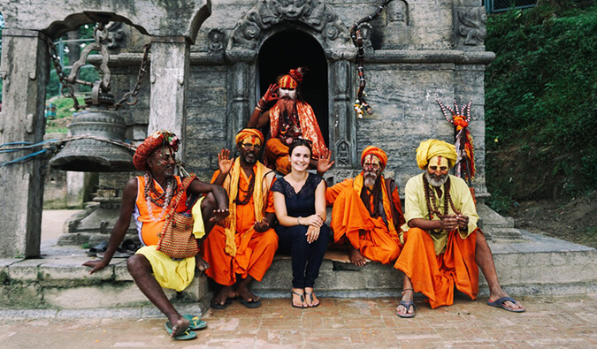 一名女子坐在印度一座寺庙外，与5名身着传统服饰、脸上涂满彩绘的男子坐在一起